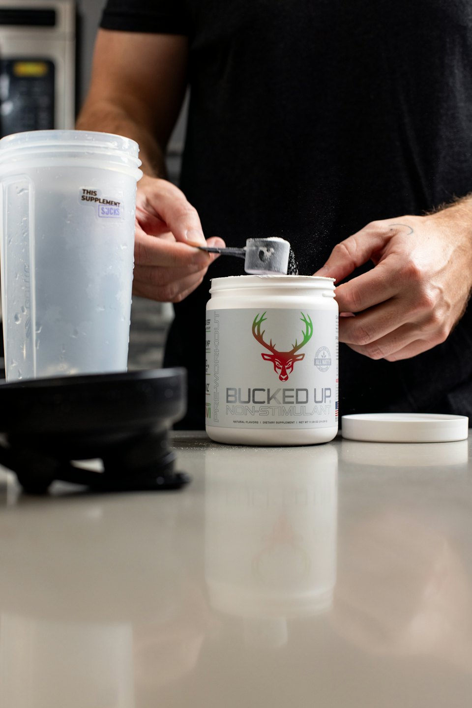 Person scoops white supplement powder from a container into a shaker.