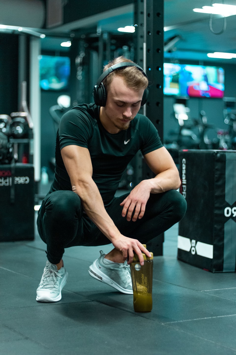 Man in athletic wear crouches in gym holding green drink.